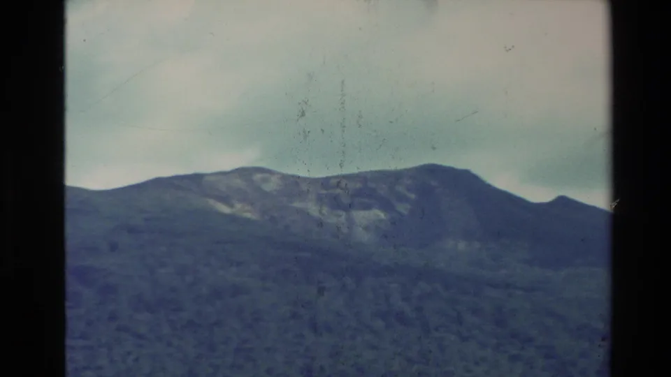 New Zealand 1981 home-movie frame showing layered mountain ridges beneath an overcast sky with visible 8mm grain and gate edge.