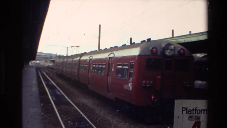 1981 New Zealand 8mm frame showing a red passenger train at a station platform, seen through dark train-window edges.