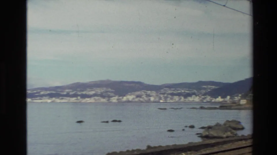 1981 New Zealand coastal scenery viewed from a moving train, with water, rocks, distant hills, and a shoreline town.