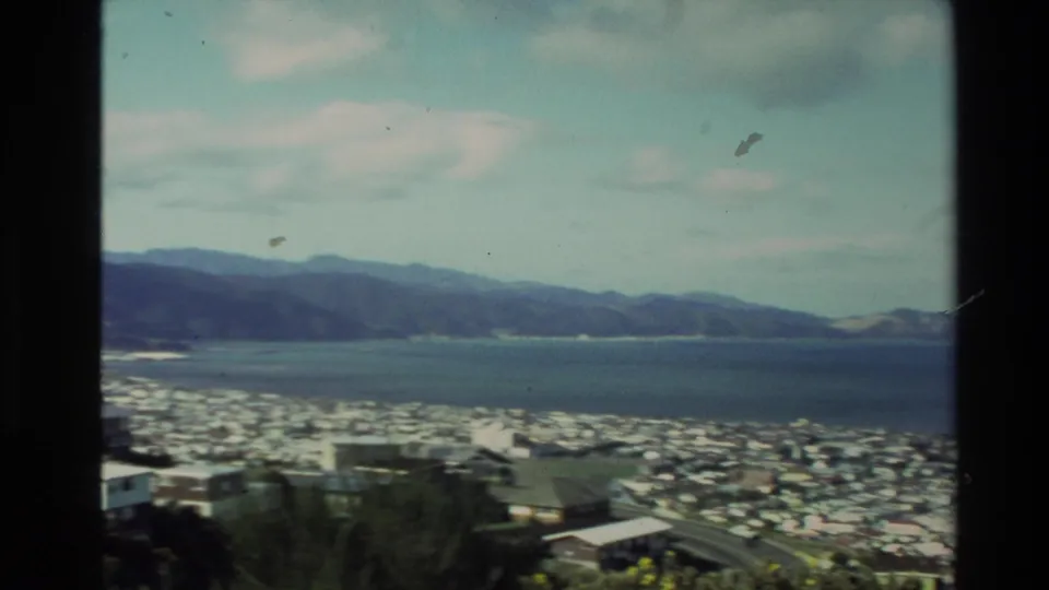 1981 New Zealand 8mm frame of a shoreline suburb beneath dark hills, with calm water stretching across the middle distance.
