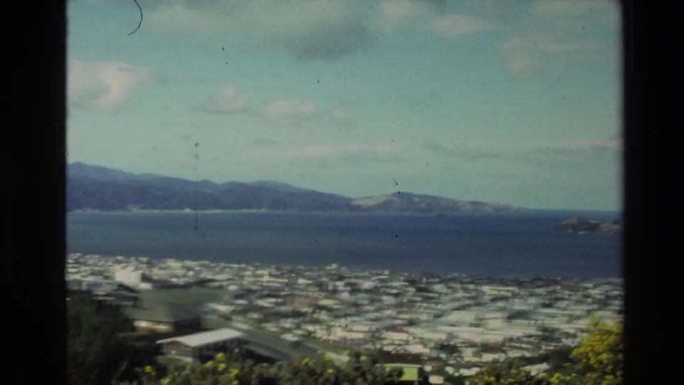 1981 New Zealand home-movie panorama of a coastal city beside a bay, with distant hills and foreground residential roofs.