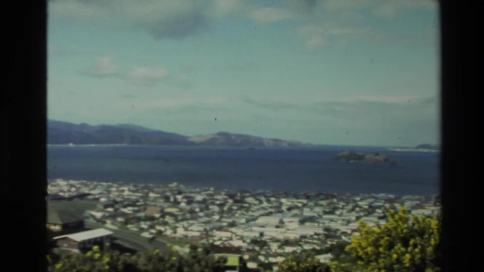 1981 New Zealand home-movie frame showing a coastal city below, harbor water, distant hills, and dark scan borders.