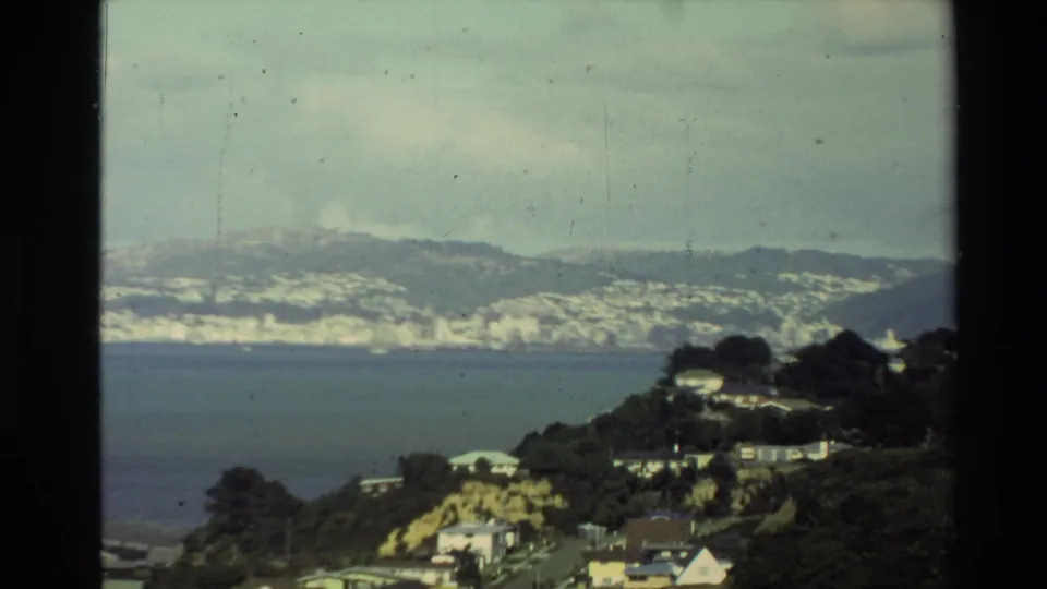1981 New Zealand home-movie frame showing a dark coastal hillside, scattered houses, bay water, and hazy mountains beyond.