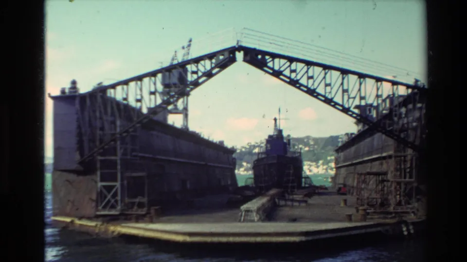 New Zealand harbor shipyard scene with a large dry dock frame, industrial buildings, water, and a vessel in the center.