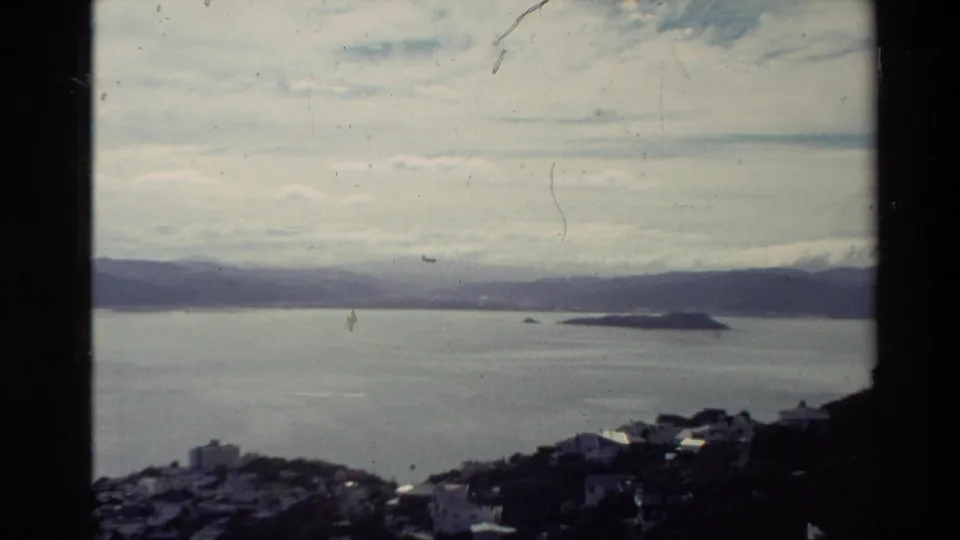 1981 New Zealand 8mm view from a hillside over blue-gray ocean, a small island, distant mountains, clouds, and dark foreground buildings.