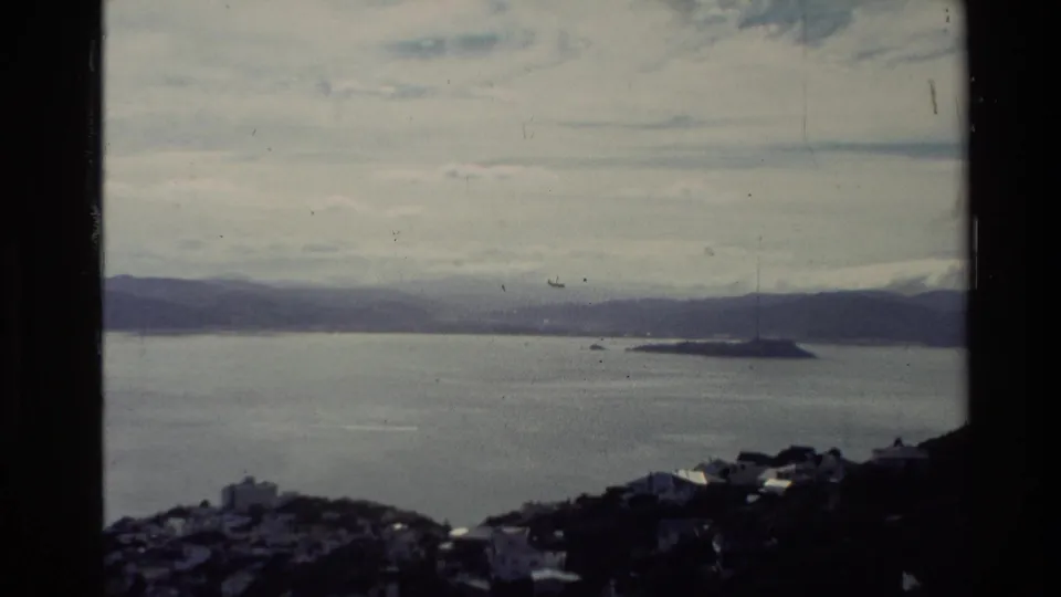 1981 New Zealand home-movie frame showing a wide coastal vista with a small island, distant hills, layered clouds, and dark foreground roofs.