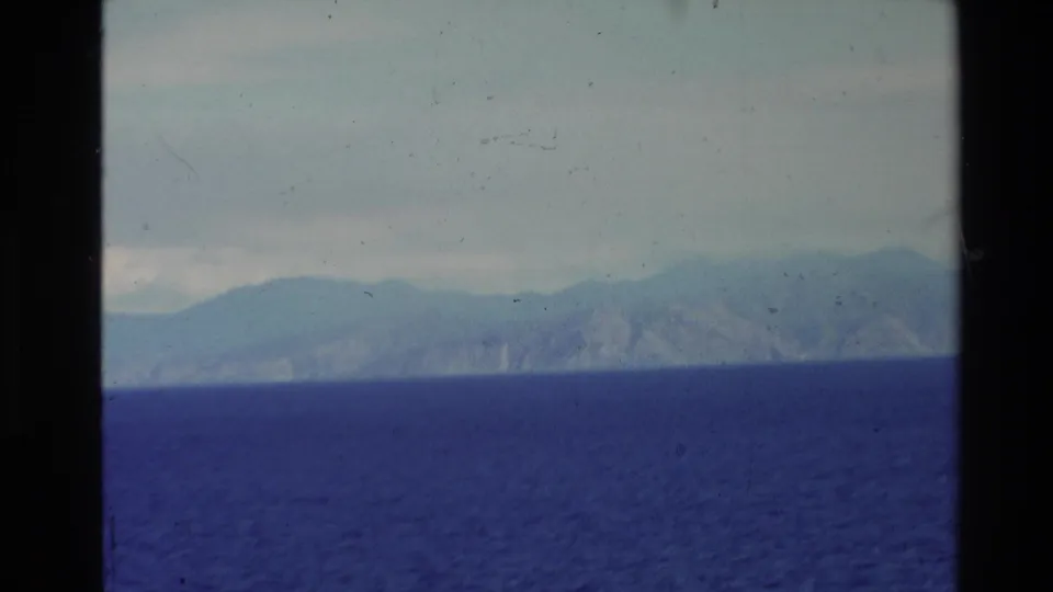 Distant New Zealand mountain coastline over deep blue sea, with soft focus, dust, and faded 8mm color.