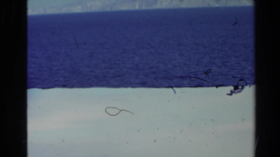 8mm travel frame from New Zealand showing open blue sea, a pale foreground band, and marked film dust and scratches.