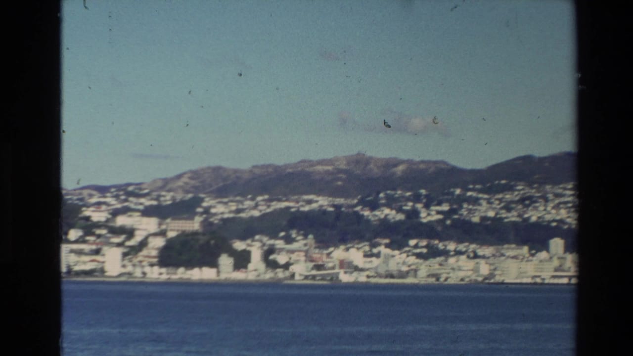 A coastal city and hills are seen across blue water from a moving vessel.