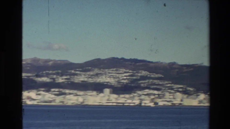 The 8mm frame shows a distant New Zealand coastal city along the waterfront, backed by hills and low mountains.
