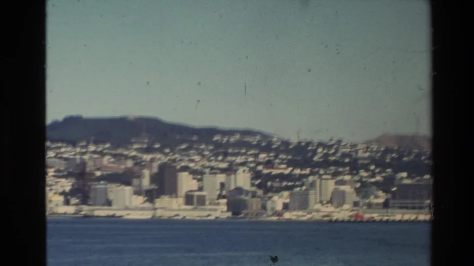 1981 New Zealand 8mm view of a waterfront city with pale buildings, hillside housing, blue water, and dark vertical frame edges.