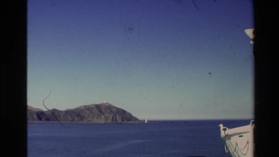 1981 New Zealand 8mm frame looking across open sea toward a low headland, with a small sail and boat structure at the right edge.