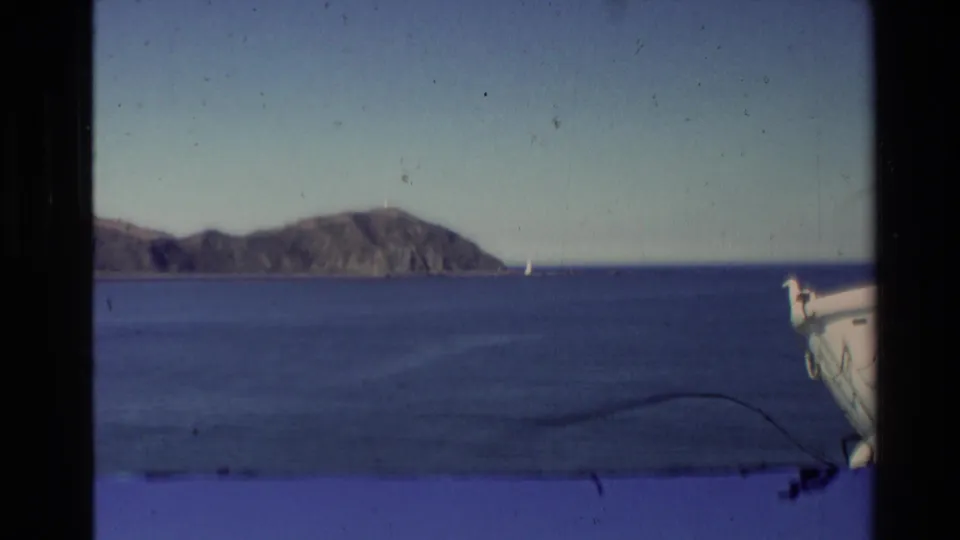 1981 New Zealand 8mm view across blue harbor water to a distant headland, with a boat edge, small sail, dust, and scratches visible.