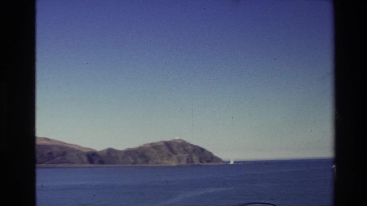 1981 New Zealand home-movie view of calm coastal water and a mountainous island under a clear sky, framed by dark window or gate edges.