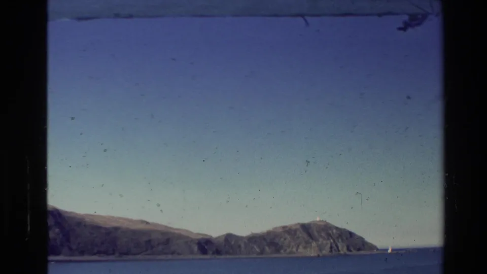 1981 New Zealand coastal landscape with a rugged island, open water, clear sky, and dark framing from a window or film gate.