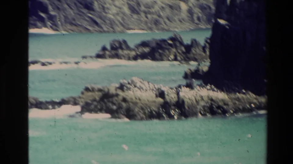1981 New Zealand home-movie frame of turquoise shallows, rocky shoreline, coastal vegetation, and steep landforms beyond the water.