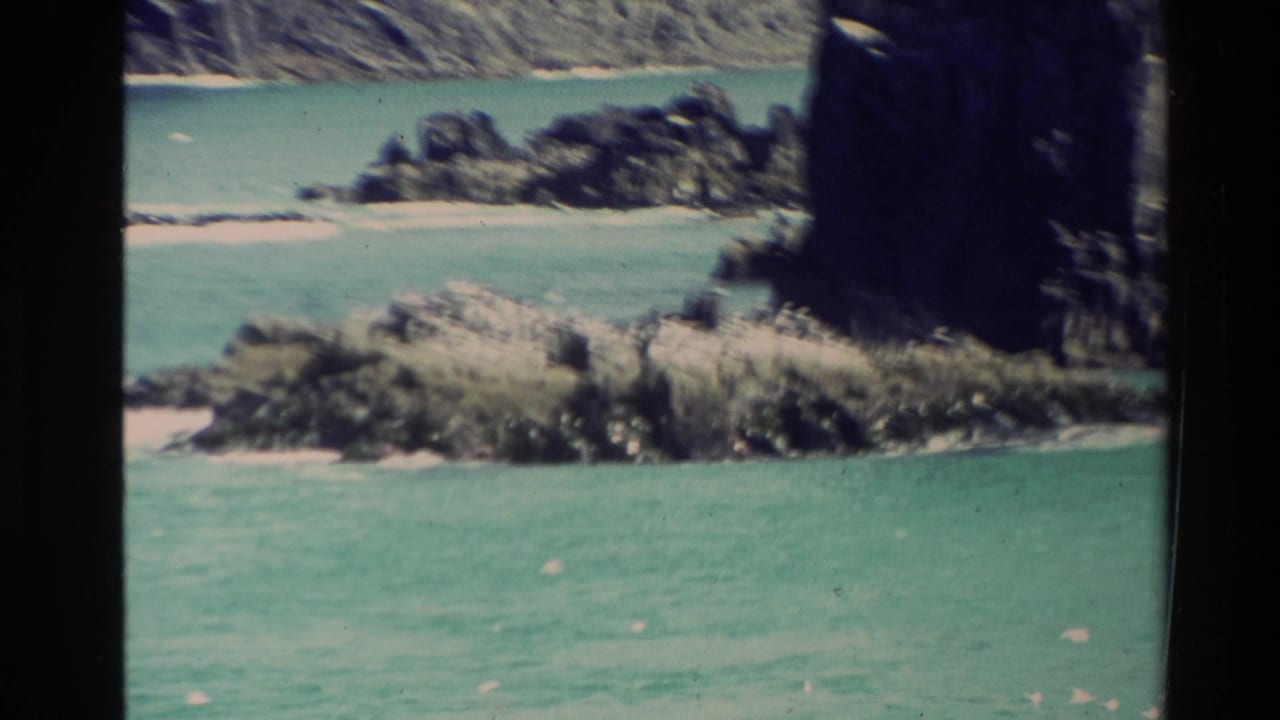 1981 New Zealand 8mm frame showing turquoise water, rugged rocks, a small sandy beach, and a dark cliff edge.
