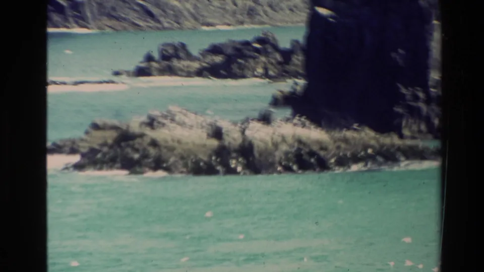 1981 New Zealand 8mm frame showing turquoise water, rugged rocks, a small sandy beach, and a dark cliff edge.