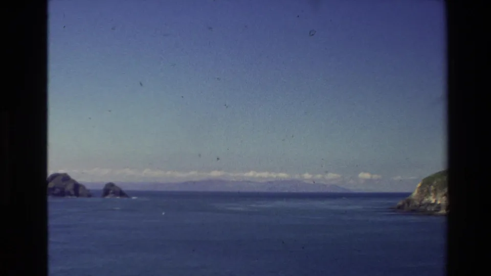 New Zealand 1981 8mm frame looking over blue coastal water toward small islands and a right-edge headland.