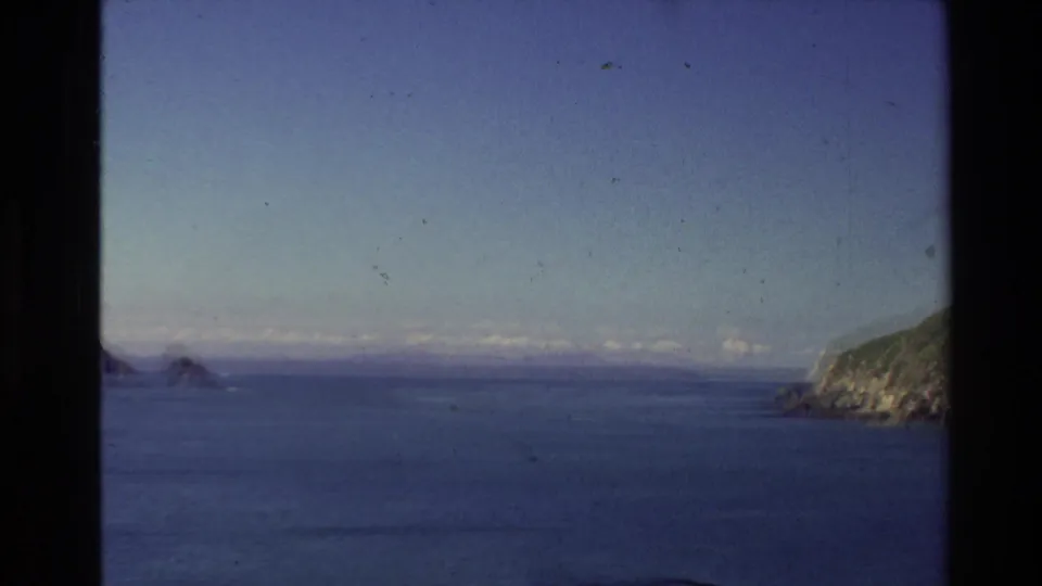 1981 New Zealand 8mm coastal view with distant island rocks, calm blue water, clear sky, and a green headland.