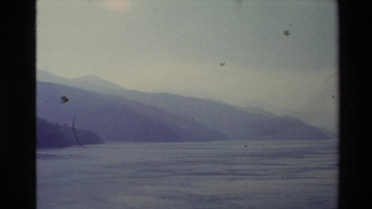 1981 New Zealand home-movie frame showing layered mountains and still lake water seen from a boat in soft, hazy light.