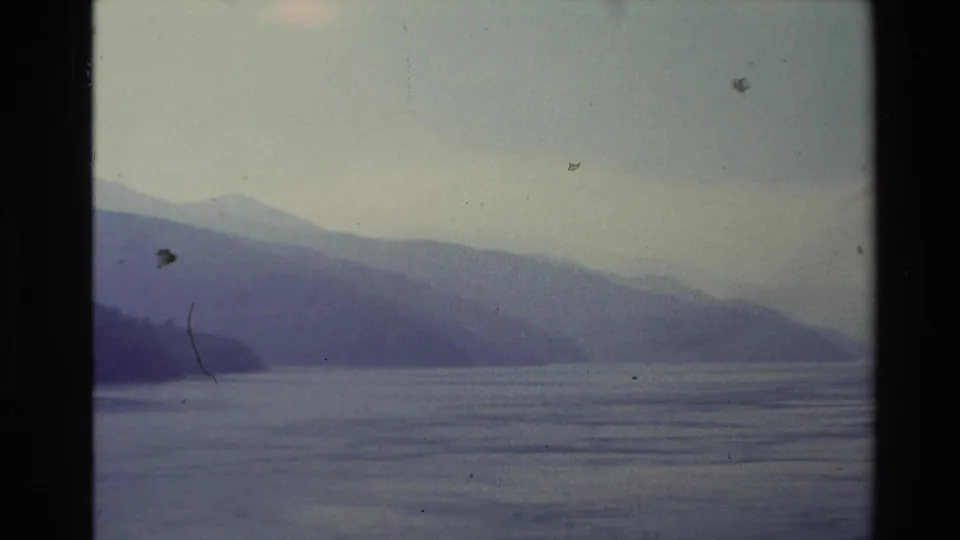 1981 New Zealand home-movie frame showing layered mountains and still lake water seen from a boat in soft, hazy light.