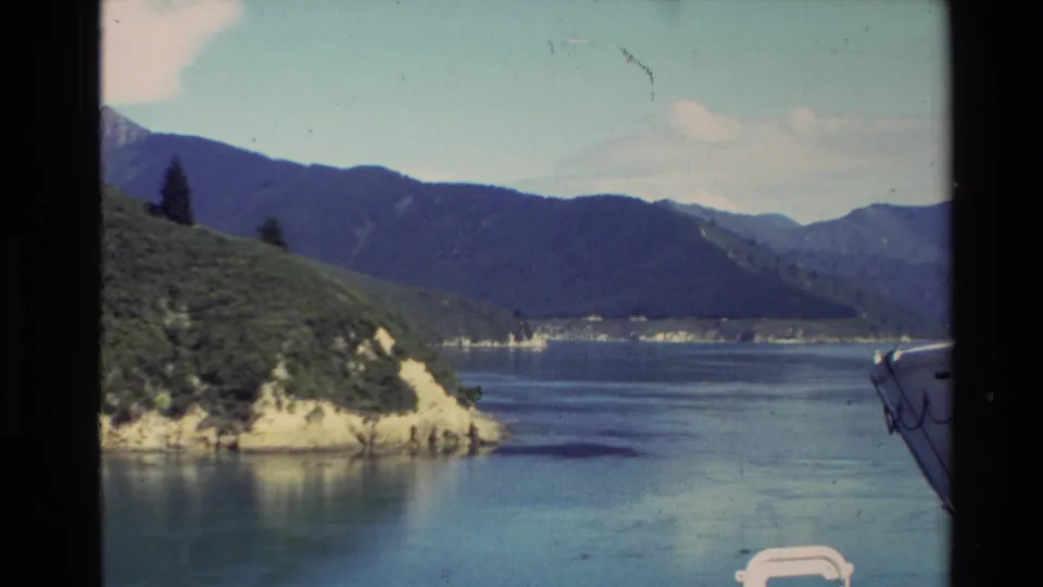 Clearer 1981 New Zealand boat-view frame showing a green headland, bright lake water, surrounding mountains, and part of the vessel edge.