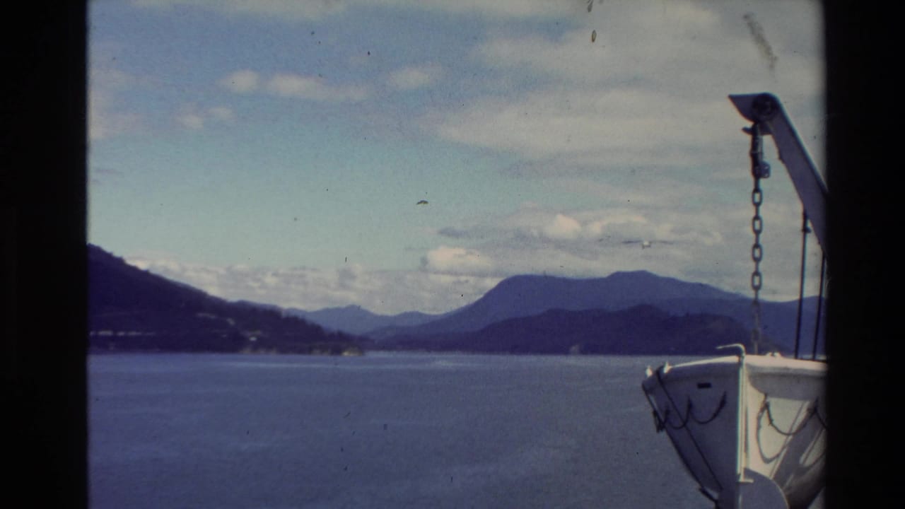 Boat-side view over a New Zealand lake, with distant mountain ridges, pale clouds, and white bow rigging at frame right.
