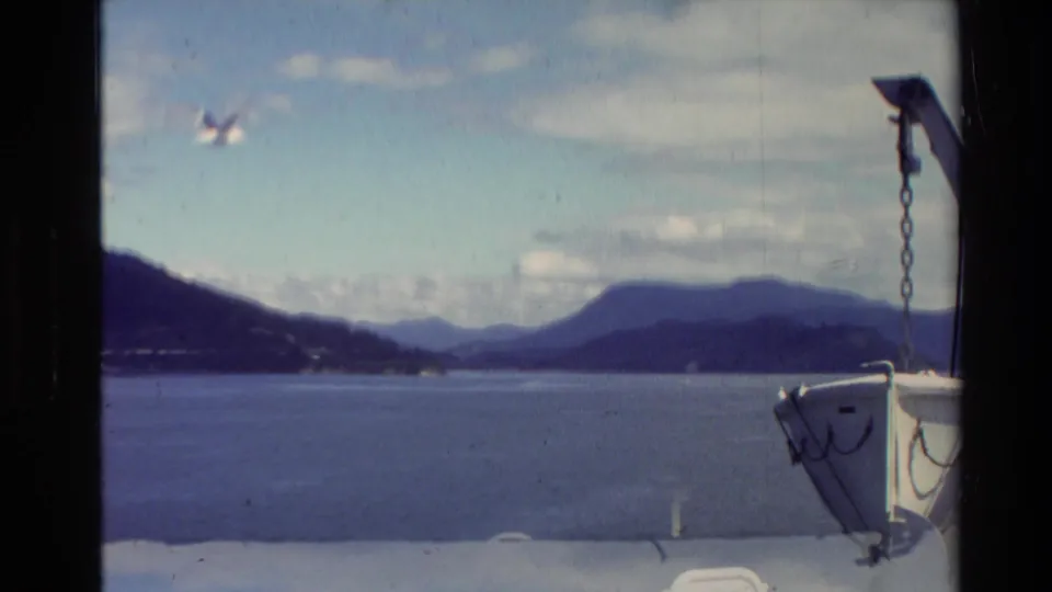 New Zealand lake landscape from a moving boat, showing the bow at right, broad water, mountain ridges, and 8mm film wear.