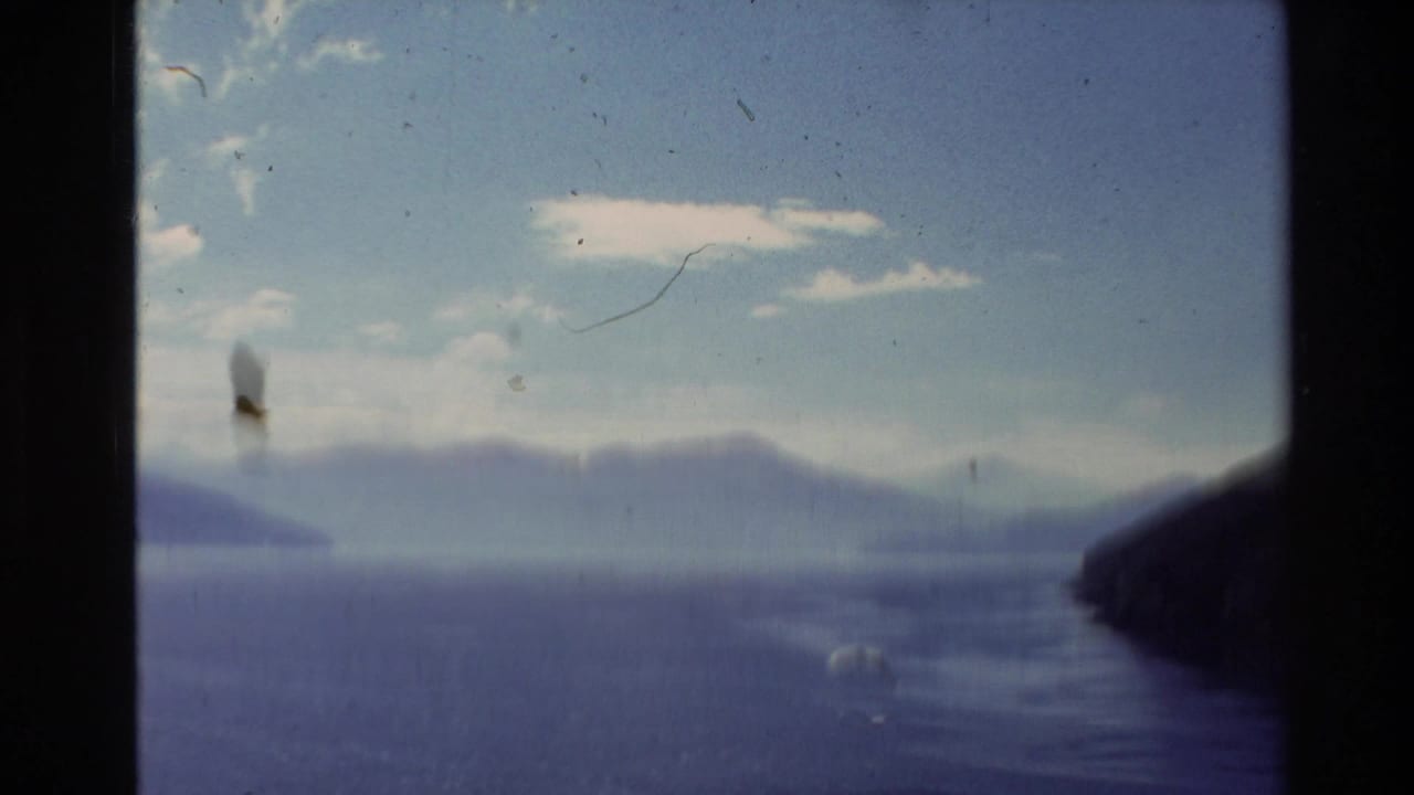 1981 New Zealand 8mm frame showing a hazy lake and distant hills beneath scattered clouds, with dark gate edges and film artifacts.