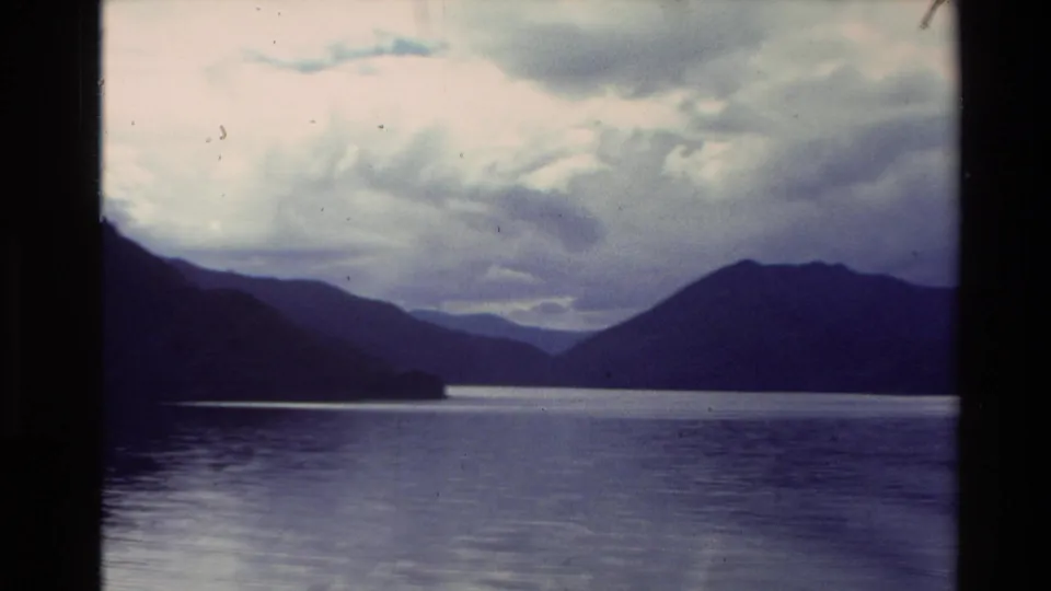 8mm travel frame from New Zealand in 1981 showing shadowed hills around a reflective lake under heavier cloud cover.