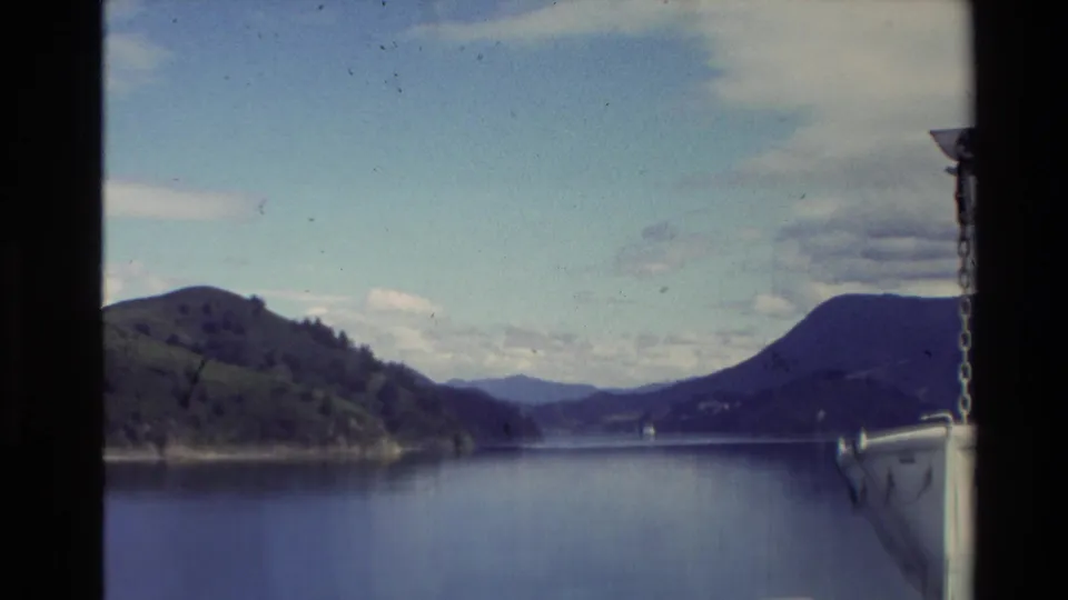 1981 New Zealand 8mm boat-view frame with calm lake water, green hillside, distant hills, and a chain visible along the right edge.