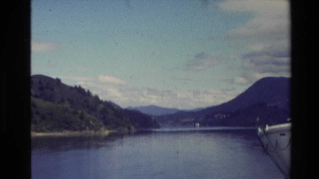 1981 New Zealand 8mm frame showing a calm lake from a boat, with wooded shoreline, rounded mountain ridges, and pale blue sky.