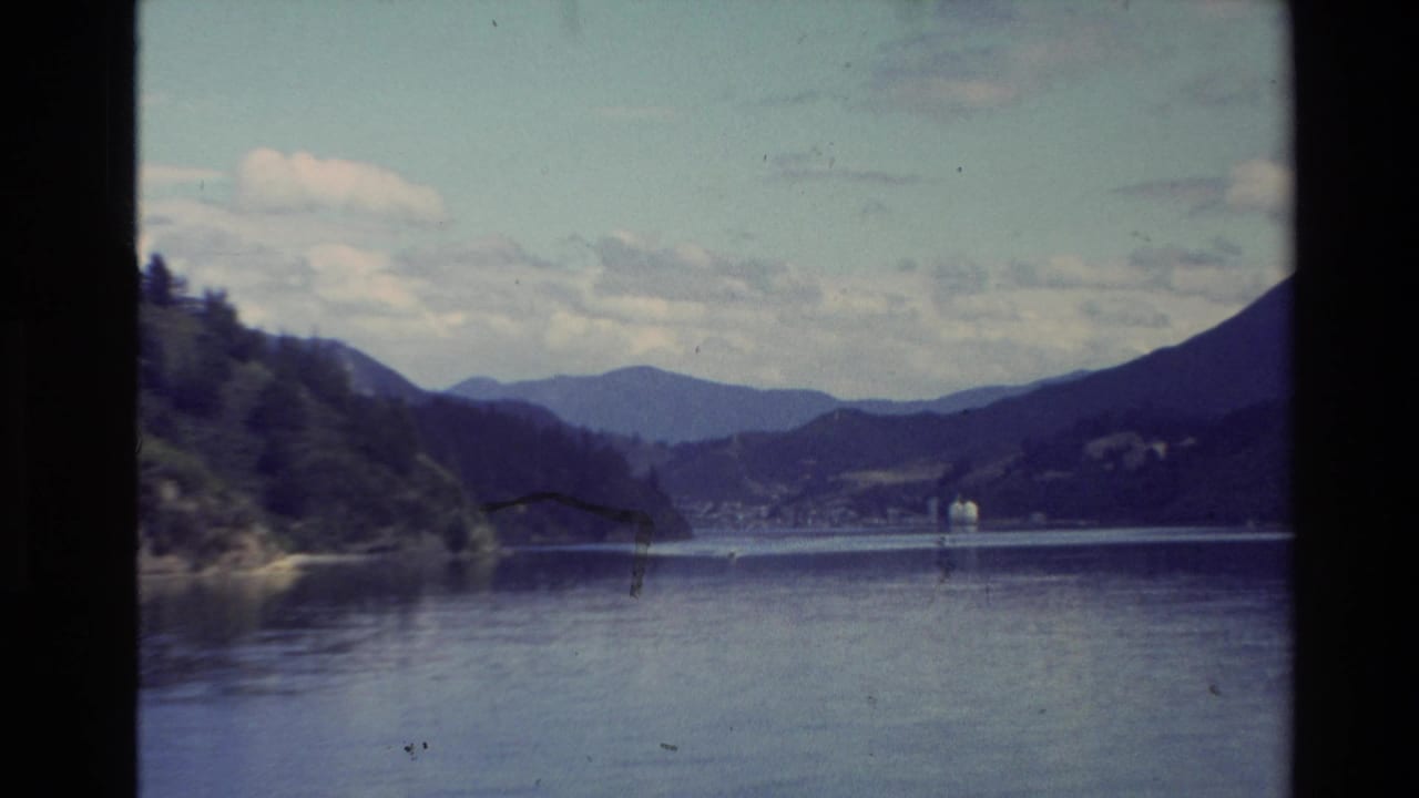 Wide lake view with wooded shoreline, dark mountain slopes, distant buildings, and blue sky.