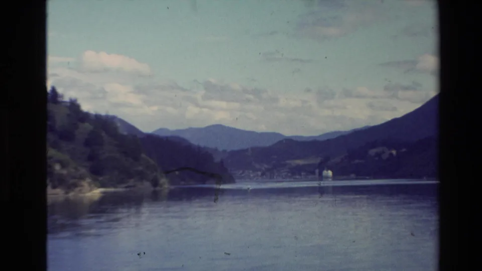 1981 New Zealand 8mm frame looking across a quiet lake toward tree-covered land, mountain ridges, and a small distant lakeside settlement.