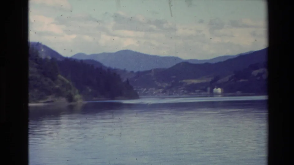 1981 New Zealand 8mm frame with calm lake water, tree-covered shoreline, layered blue mountain ridges, and distant lakeside buildings.