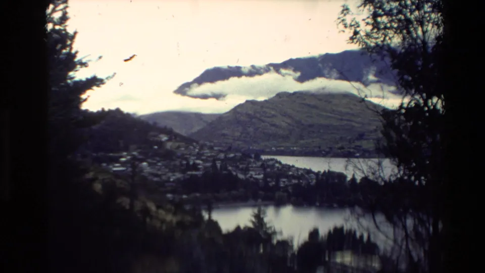1981 New Zealand 8mm frame from a hillside or overlook showing a lakeside town, water, dark trees, mountains, and a low cloud.