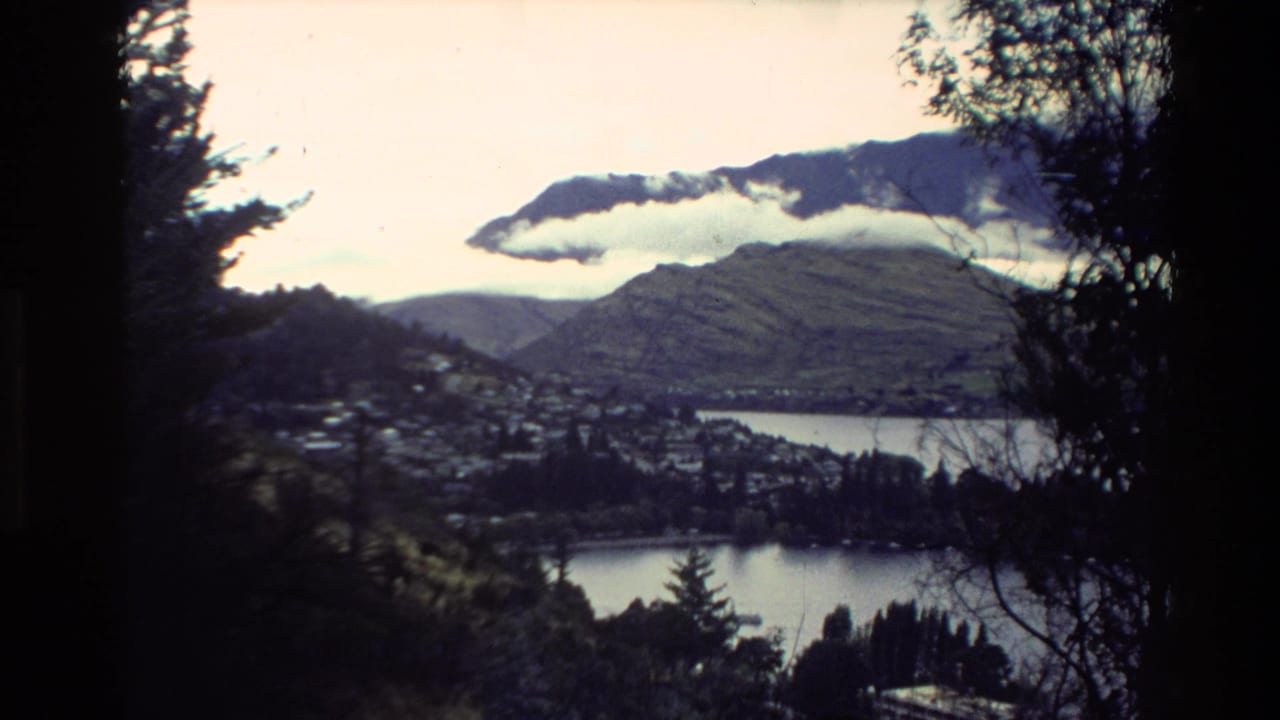 1981 New Zealand home-movie view of a lakeside town and mountains seen through dark tree silhouettes under low cloud.