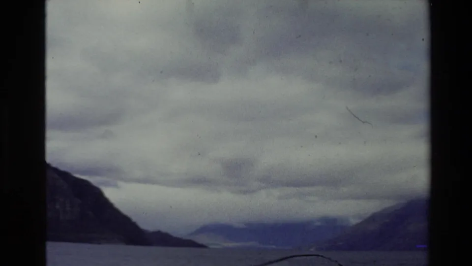 Vintage New Zealand travel footage shows a broad lake or fjord-like waterway between mountains under layered cloud.