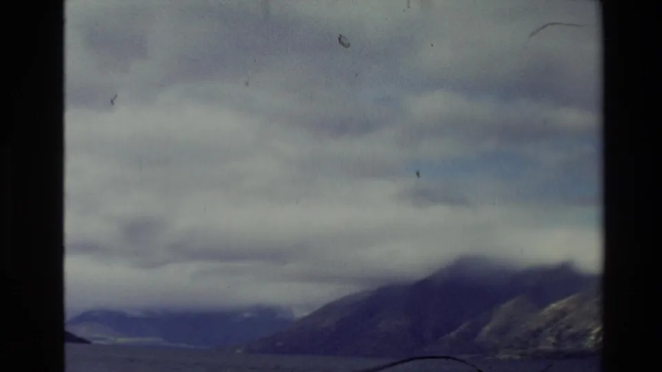 1981 New Zealand home-movie frame of a cloudy mountain lake view, with slopes enclosing the water and visible 8mm grain.