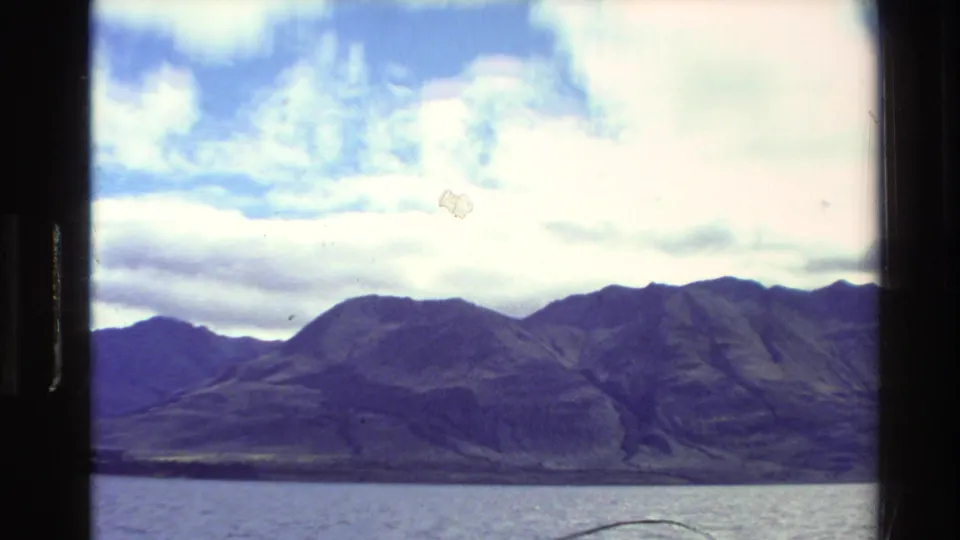 1981 New Zealand 8mm frame showing mountains across water under bright clouds, with grain and minor scan marks.