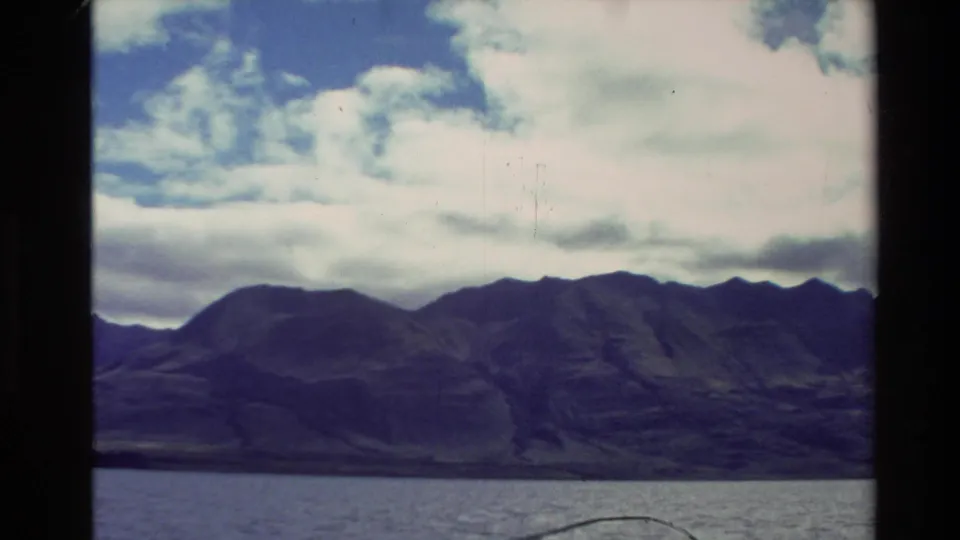 1981 New Zealand 8mm frame showing a dark mountain range beyond a calm lake under broken cloud.