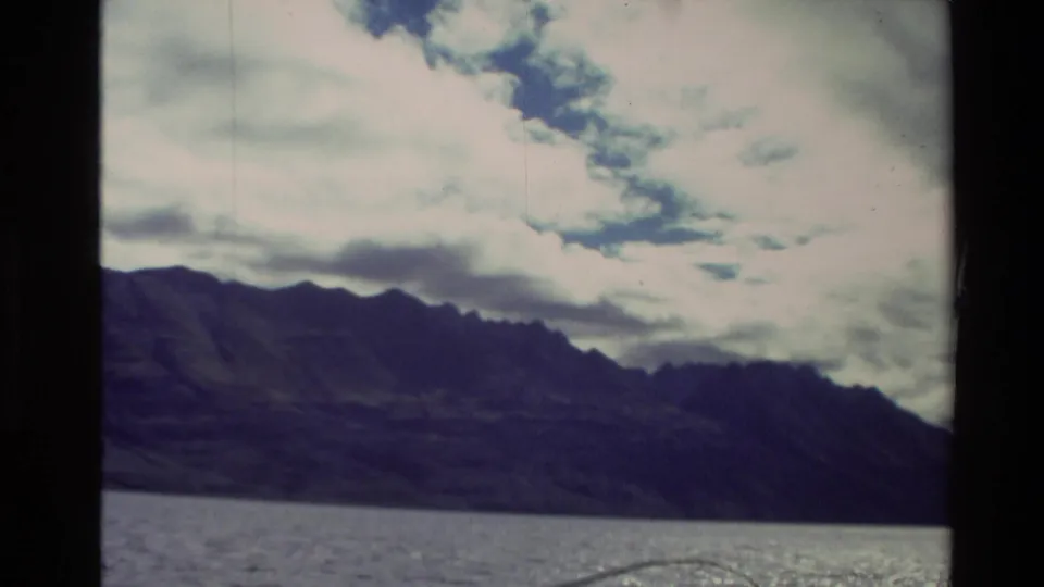 New Zealand 8mm travel frame from 1981 showing lake water, shadowed mountain slopes, and bright clouds.