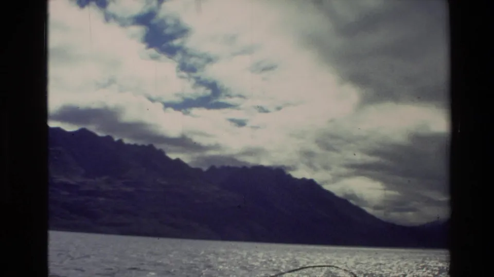 1981 New Zealand 8mm frame with sun glinting on lake water below a dark mountain ridge and heavy cloud.