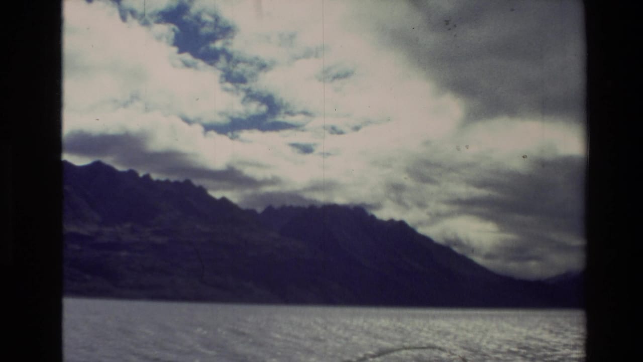 1981 New Zealand 8mm frame showing a dark mountain ridge beyond a lake under heavy cloud, with visible film border and grain.