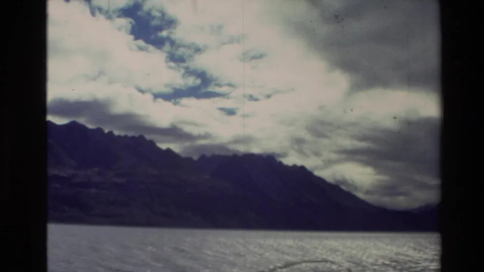 1981 New Zealand 8mm frame showing a dark mountain ridge beyond a lake under heavy cloud, with visible film border and grain.