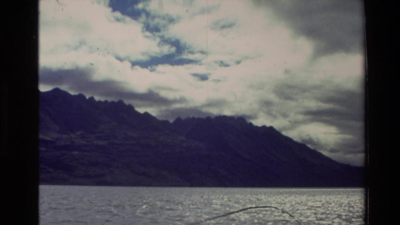 1981 New Zealand 8mm frame with rugged mountains across a lake, bright cloud cover, and a strong vertical film divider.