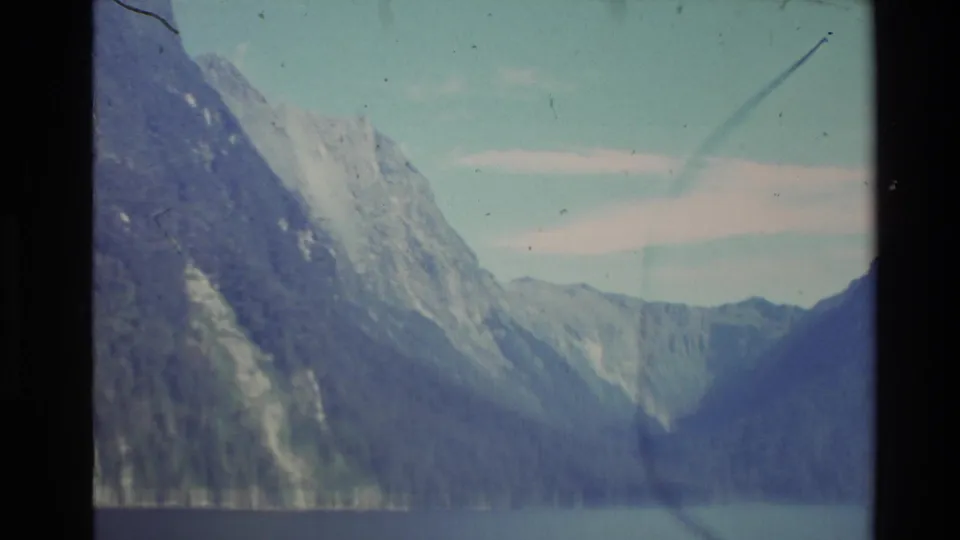 1981 New Zealand 8mm frame showing pale alpine slopes above water, with faded color, soft focus, and handheld composition.