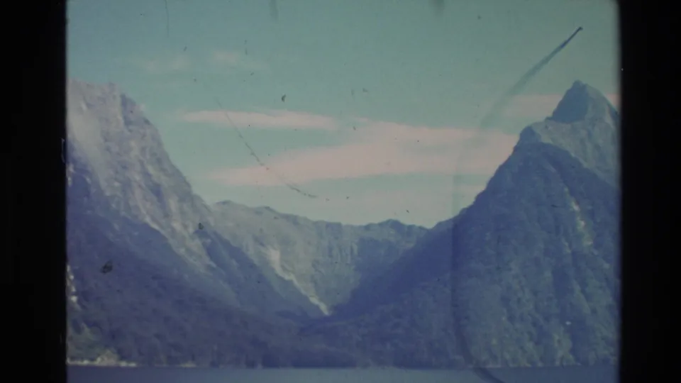 1981 New Zealand 8mm frame with steep mountain walls, a lake foreground, pale sky, and visible scratches from the film scan.