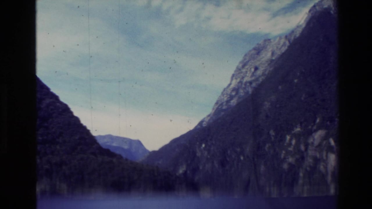 Vintage 8mm view of a New Zealand fjord corridor, with dark steep slopes and blue water receding toward distant hills.